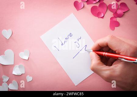 Hand writing I love you on white sheet on pink background with pink and white paper heart cutouts. Top view. Stock Photo