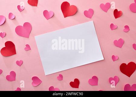 Background with cutouts of pink and red paper hearts on pink with empty white card in the center. Top view. Stock Photo