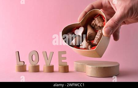 Hand offering box of heart shaped chocolates on pink background with wooden letters with word love. Front view. Stock Photo