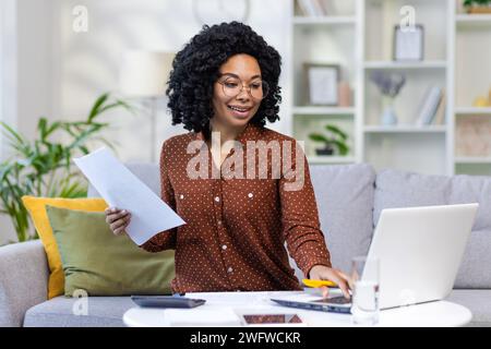 Young smiling African American woman working from home with documents and bills, sitting on sofa at home and using laptop. Stock Photo