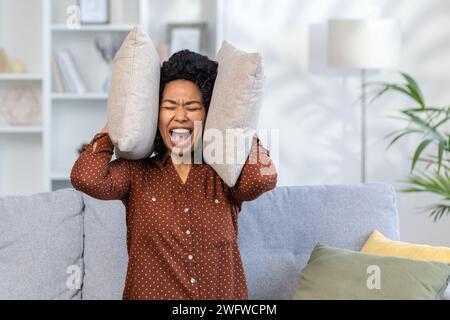 Close-up photo of a young African-American woman sitting on the couch at home and hoarsely screaming with her eyes closed and pillows covering her ears. Stock Photo
