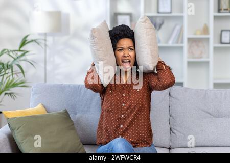 Dissatisfied and angry African American young woman sitting on sofa at home and covering ears with pillows from excessive noise. Stock Photo