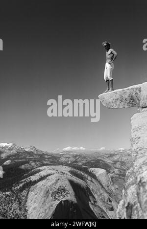 Young man risks his life and stands on a rocky ledge on the summit of Half Dome, Yosemite Valley, California, USA Stock Photo