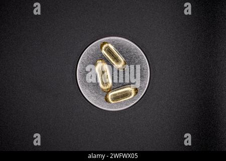 Omega 3 fish oil gel capsules placed on an aluminum cap on a neutral black background. Cod liver tablets. Three of high contrast omega 3-6-9. Stock Photo