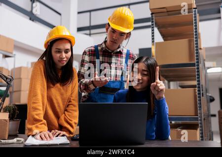 Asian delivery service managers analyzing logistics on laptop while standing near desk in warehouse. Storehouse package handlers and shipment operator looking at orders checklist on computer Stock Photo