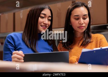Storehouse asian women managers discussing goods stocking operations while standing near shelf full of cardboard boxes. Warehouse order pickers comparing customer checklist and pick ticket Stock Photo