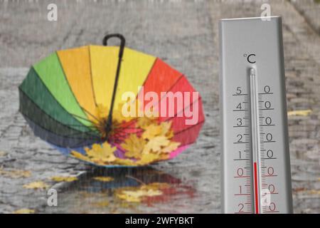 Thermometer and open colorful umbrella with fallen leaves on wet pavement under rain. Temperature in autumn Stock Photo