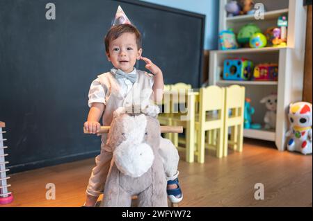 Cute one year old multiracial baby boy celebrating his first birthday in kindergarten. He is riding a toy wooden horse and smiling sweetly Stock Photo