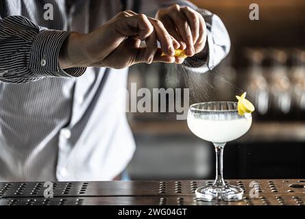 A bartender squeezes a lemon peel and squirts the juice onto a French 75 cocktail drink on the bar counter. Stock Photo