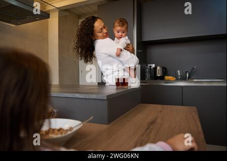 Selective focus of a beautiful young mother holding baby boy son while older daughter sitting near bowl with delicious flakes, enjoying happy time tog Stock Photo