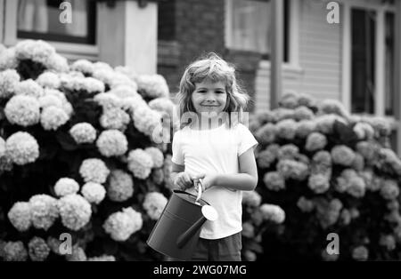 Cute little boy watering flowers with watering can in the garden. Child dressed in light summer closes and colourful t-shirt, smiling and having fun. Stock Photo