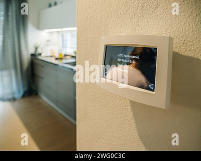 Modern smart home thermostat displaying temperature settings on a wall with blurred kitchen background. Stock Photo