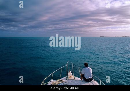 A man sits on the bow of a boat crossing a shallow lagoon. Stock Photo