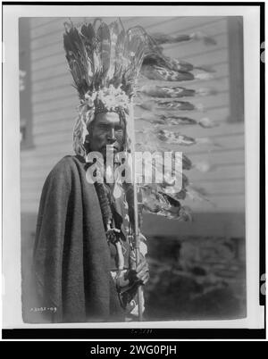 Piegan war-bonnet and coup-stick, c1910. Piegan man, half-length portrait, dressed in war bonnet and holding a feathered coup-stick, facing front. Stock Photo