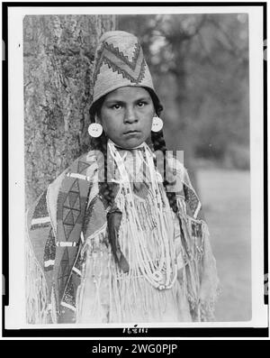 Umatilla maiden, c1910. Umatilla woman, half-length portrait, facing front, standing in front of tree, wearing beaded buckskin dress, shell bead necklaces, shell disk earrings, and woven grass hat. Stock Photo