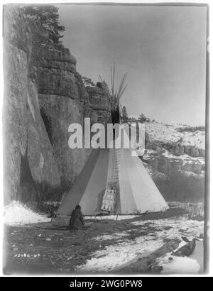 Hide stretching-Apsaroke, c1908. Apsaroke woman stretching hide that has been secured to the ground by stakes, tipi in background, cliff on left. Stock Photo