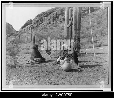 Qahatika women resting in Harvest Field-Qahatika, c1907. Three Qahatika women sitting on ground with kiho carriers and pot nearby, saguaro cacti and mountains in background. Stock Photo