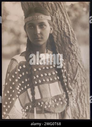 Lucille [B], c1907. Photograph shows Lucille, a Native woman, half-length portrait, standing next to tree, facing front, wearing a headband and seashell decorated buckskin dress. Stock Photo