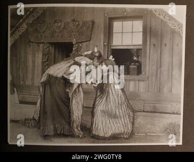 Maoris rubbing noses (Te Hongi) 1891. Two Maori girls greet each other by rubbing noses in front of a Maori house with carved decorations. Both have feathers in their long hair and wear stiff cloaks decorated with dangling cords. One wears a European dress beneath. Full length portrait. From Charles Appleton Longfellow's trip to New Zealand in 1891. Stock Photo