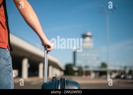 Travel by airplane. Traveler is walking from parking lot to airport terminal. Man with suitcase against air traffic control tower on sunny summer day. Stock Photo