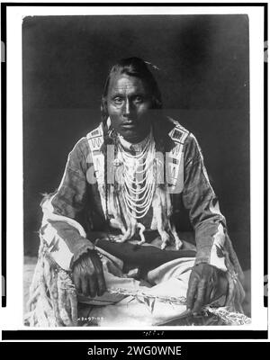 Wades in Water, Piegan Indian, full-length portrait, seated on floor, facing front, with braids, beaded buckskin shirt and leggings, beads with ermine tail trim, c1910. Stock Photo