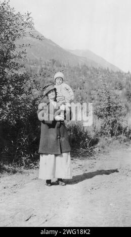 Woman standing on dirt road holding child; mountains in background, between c1900 and 1916. Stock Photo