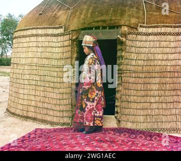 Full-length profile portrait of a woman, possibly Turkman or Kirgiz, standing on a carpet at the entrance to a yurt, dressed in traditional clothing and jewelry, between 1905 and 1915. Ethnic Diversity: Profile of a Nomad. In this portrait, Prokudin-Gorksii captures the traditional dress, jewelry, and hairstyle of an Uzbek woman. A yurt is a portable tent used for housing by the nomadic peoples of Central Asia. After conquering Turkestan in the mid 1800s, the Russian government exerted strong pressure on the nomadic peoples to adopt a sedentary lifestyle and settle permanently in villages, tow Stock Photo
