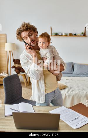 single father holding infant son while talking on smartphone near laptop on desk, remote work Stock Photo