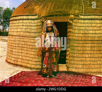 Full-length profile portrait of a woman, possibly Turkman or Kirgiz, standing on a carpet at the entrance to a yurt, dressed in traditional clothing and jewelry, between 1905 and 1915. Ethnic Diversity: Profile of a Nomad. In this portrait, Prokudin-Gorksii captures the traditional dress, jewelry, and hairstyle of an Uzbek woman. A yurt is a portable tent used for housing by the nomadic peoples of Central Asia. After conquering Turkestan in the mid 1800s, the Russian government exerted strong pressure on the nomadic peoples to adopt a sedentary lifestyle and settle permanently in villages, tow Stock Photo
