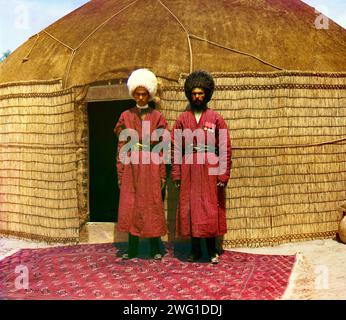 Two men standing on a rug, in front of yurt, between 1905 and 1915. Portrait of two men, possibly Turkman or Kirgiz, in traditional dress. A yurt is a portable tent used for housing by the nomadic peoples of Central Asia. After conquering Turkestan in the mid 1800s, the Russian government exerted strong pressure on the nomadic peoples to adopt a sedentary lifestyle and settle permanently in villages, towns, and cities. Stock Photo