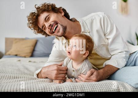happy man with curly hair laughing while embracing his baby boy and lying together on bed Stock Photo