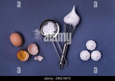 top view on uniform gray background, egg white in shell, sieve with powdered sugar, a whisk with whipped egg white and some meringues Stock Photo