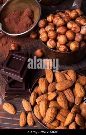 Still life. in the foreground, vintage metal measuring spoons with shelled hazelnuts and almonds, cocoa powder and pieces of dark chocolate. Stock Photo