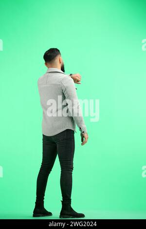 A man, full-length, on a green background, looks at his watch Stock Photo