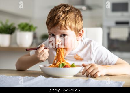 A cute little boy is eating spaghetti bolognese for lunch in the kitchen at home and is covered in ketchup. Stock Photo