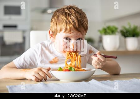 A cute little boy is eating spaghetti bolognese for lunch in the kitchen at home and is covered in ketchup. Stock Photo