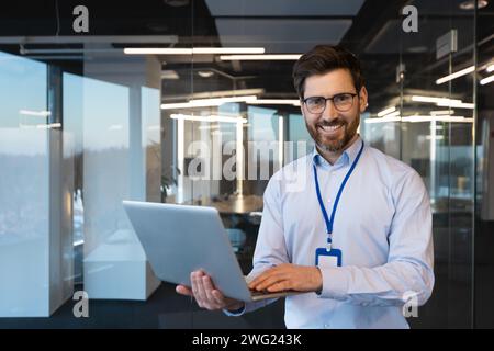 Portrait of a smiling young office worker, a specialist businessman standing in the office by the window in a shirt and wearing a badge, holding a laptop and confidently looking at the camera. Stock Photo