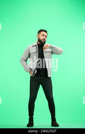 A man, full-length, on a green background, looks at his watch Stock Photo