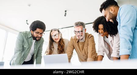 A diverse team of colleagues enthusiastically engages with content on a laptop, capturing a collaborative work culture and an effective brainstorming session in a contemporary office Stock Photo