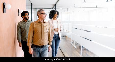 Three colleagues walk through their office space, engaged in a casual conversation that reflects a culture of openness and ongoing collaboration, highlighted by the modern and transparent design Stock Photo