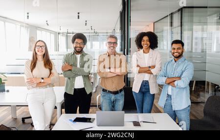 A diverse group of five colleagues stands confidently in the office, arms crossed, with smiles, symbolizing united and cooperative team ready to tackle business challenges in a co-working environment Stock Photo