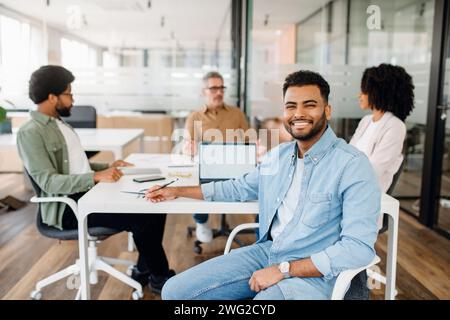 The focus is on a smiling young Arab professional in a denim shirt, sitting in front of a modern office space where his team is actively engaged in a meeting. Teamwork and brainstorming concept Stock Photo