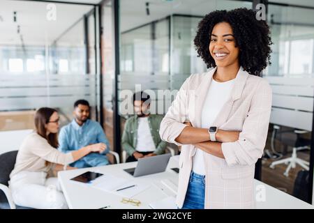 Smart high-skilled young Brazilian woman with a cheerful smile stands with arms crossed on the forefront in a modern office setting with her colleagues collaborating in the background, team synergy Stock Photo