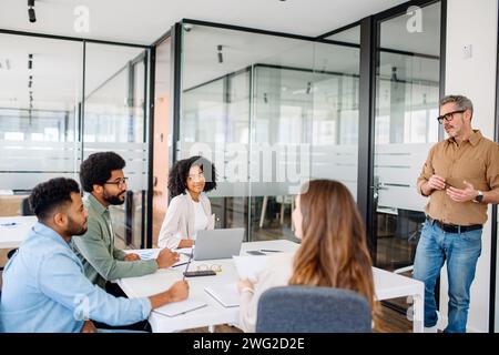 In a modern office, a man in a tan shirt gestures during a dynamic presentation to attentive colleagues around a table, illustrating a collaborative work culture and the exchange of innovative ideas Stock Photo