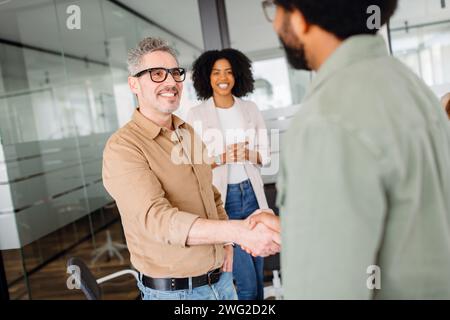 A smiling man in a tan shirt firmly shakes hands with a colleague in a green shirt, symbolizing a successful agreement or introduction, showcasing a friendly and collaborative office environment Stock Photo
