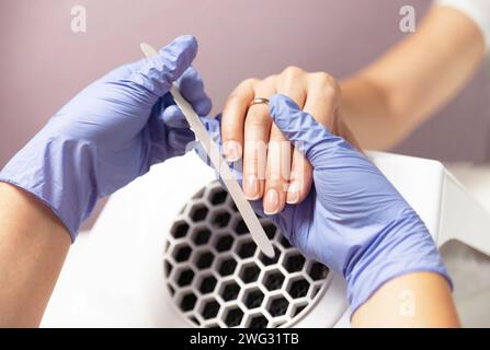 Manicure process. A master in blue gloves polishes a client's nails with a nail file in a beauty salon. Close-up. Stock Photo