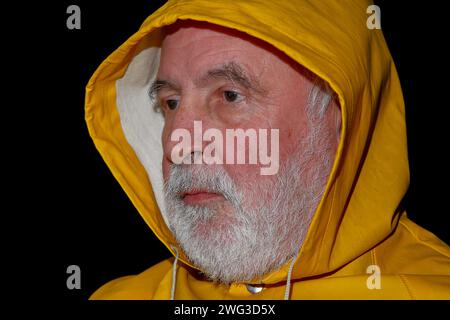 Under the hood of the yellow oil jacket, the sweat glistens on the face of the old, bearded fisherman, who looks seriously ahead of him. Stock Photo