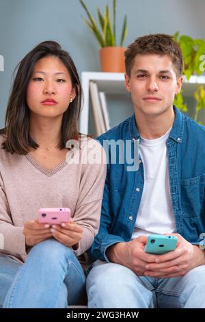 Vertical portrait of young serious multiethnic couple using smartphone technology.Indoors Stock Photo