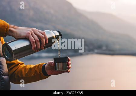 Man pouring hot tea from thermo flask with a beautiful scenic view of the mountain lake on the background Stock Photo