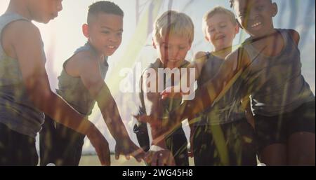 Image of sun and grasses over happy diverse schoolchildren stacking hands in outdoor sport class Stock Photo
